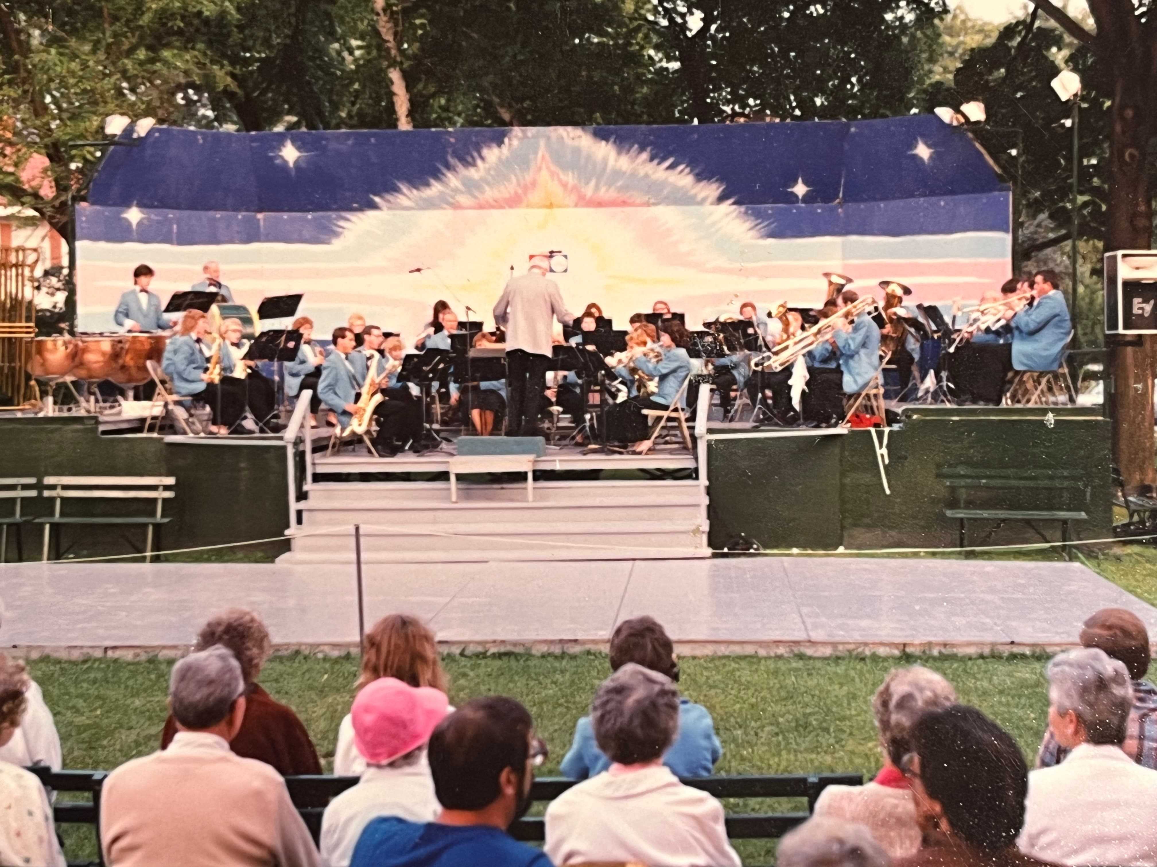 Clare Hounsell conducting the band on an old multi-colored band shell. The band is wearing light-blue suit jackets.