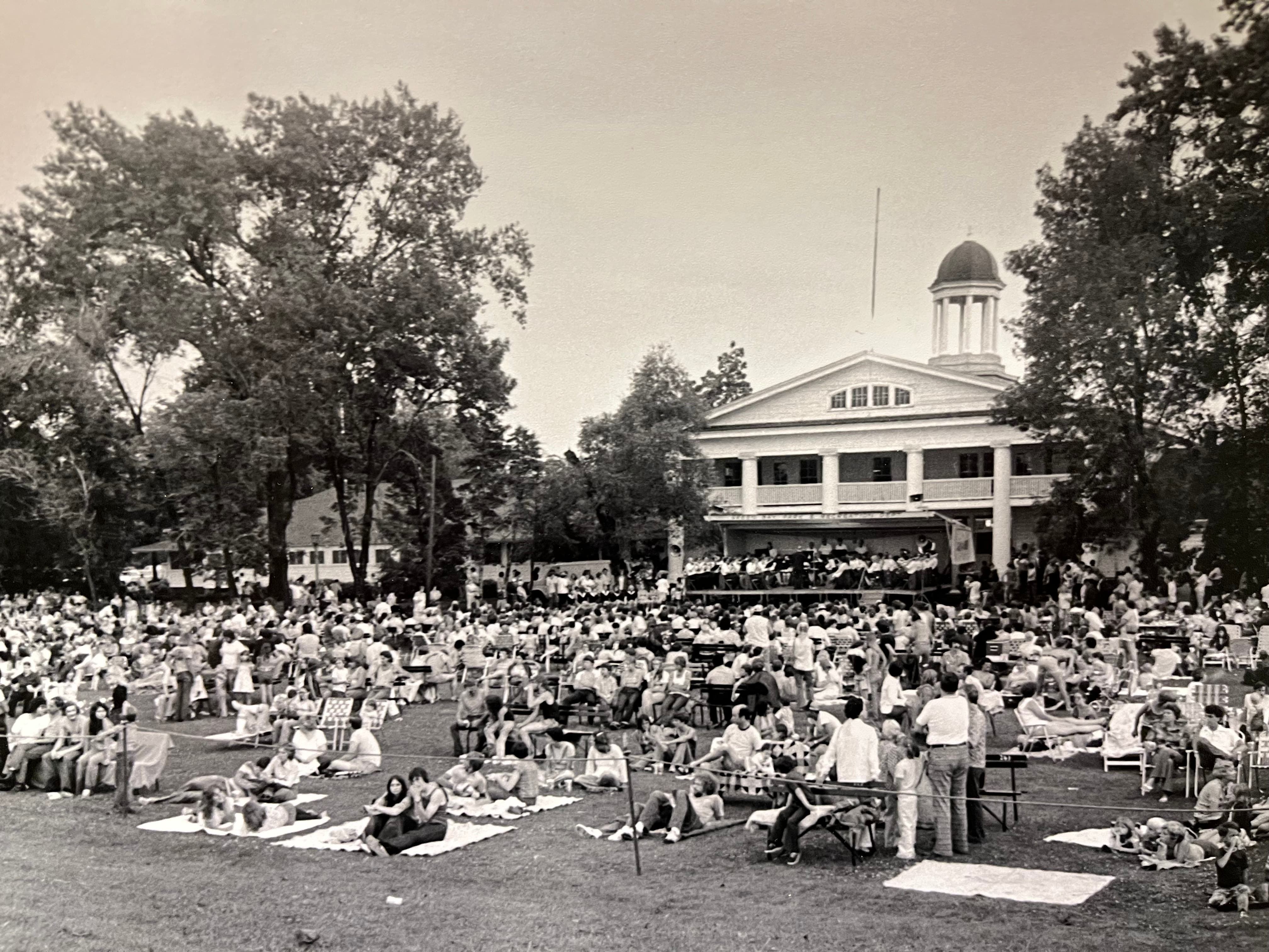A black and white photo of the band playing a concert at Bay Beach. The lawn is full of people sitting on blankets and folding chairs.